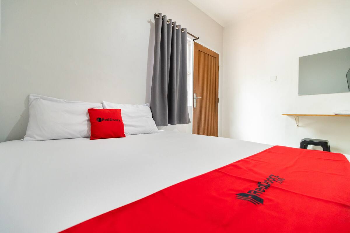 A hotel room with a bed, a red pillow, a grey curtain, a wooden door, and a small table.