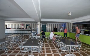 A cafeteria with metal tables and chairs, a kitchen area, and a patterned wall.