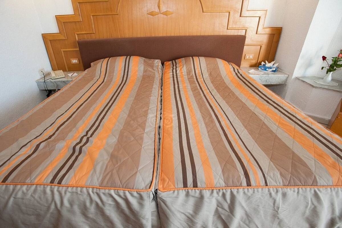 A neatly made bed with striped bedding, a wooden headboard, and a bedside table with a tissue box and a vase with a single flower.