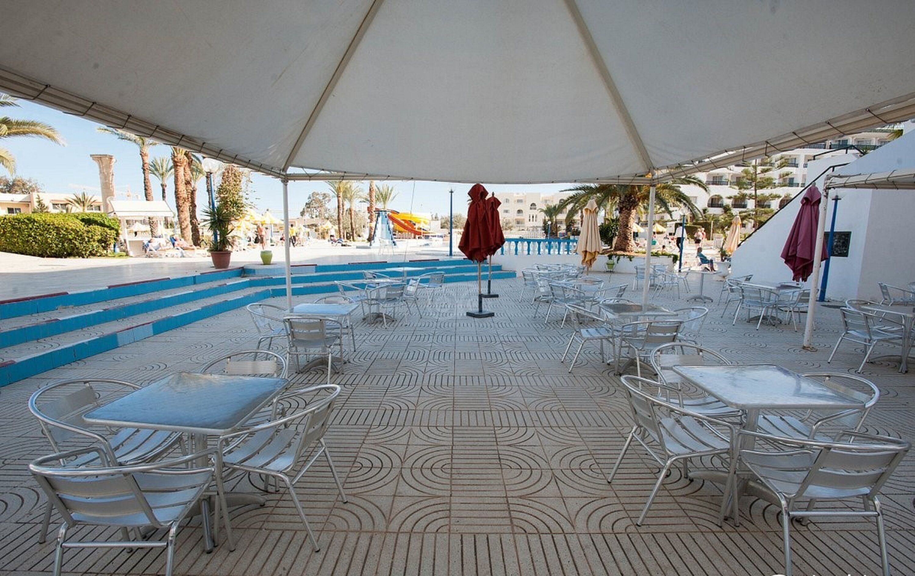 Eine Außenterrasse mit Metalltischen und -stühlen, ein überdachter Bereich mit weißem Baldachin und ein Pool im Hintergrund.