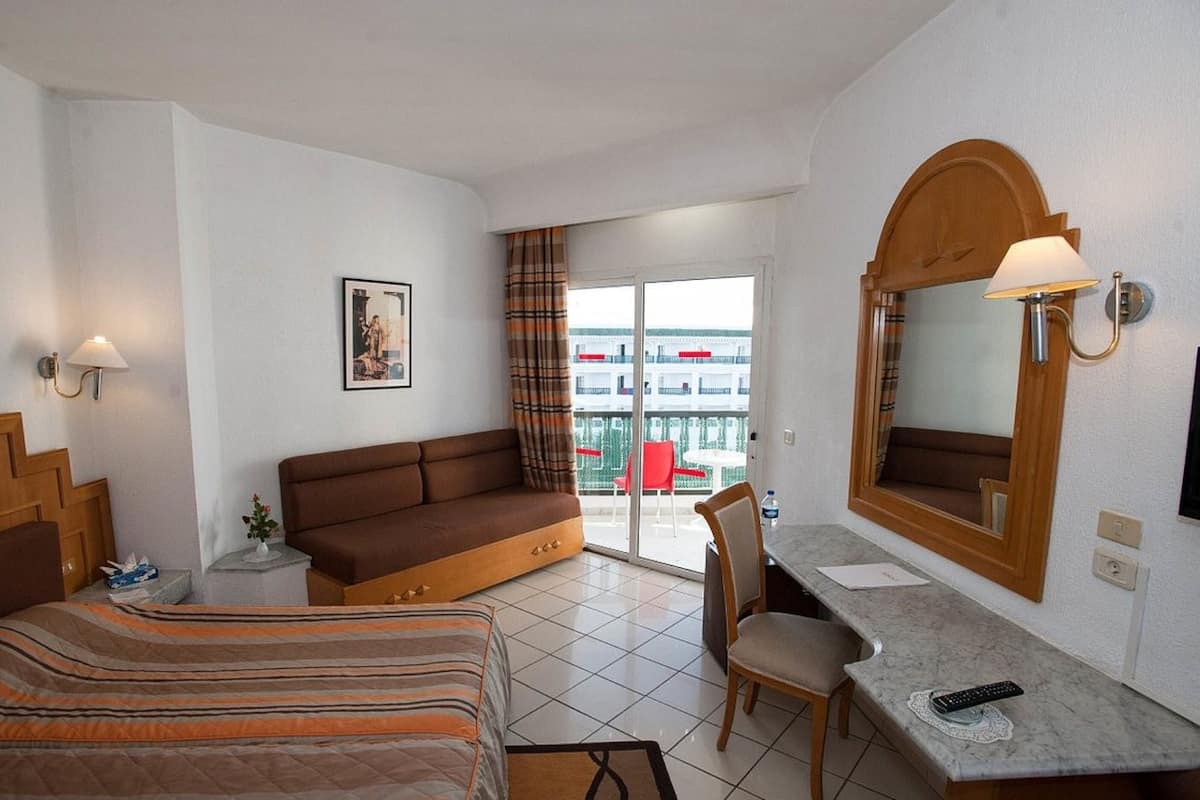 A hotel room with a bed, a sofa, a desk with a chair, a bathroom mirror, and a view of the outside balcony.