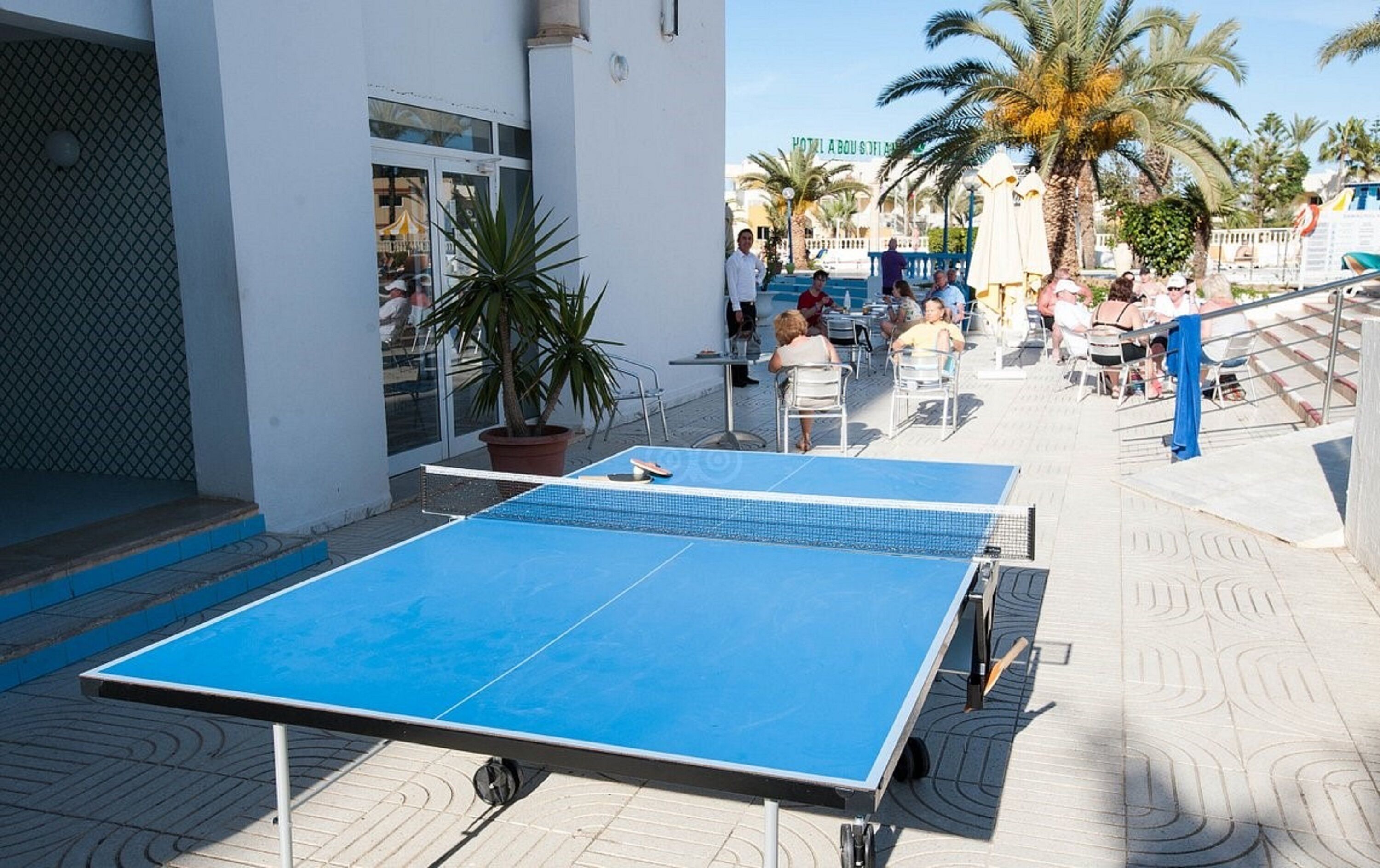 Un espace extérieur aménagé avec une table de ping-pong, avec une entrée de bâtiment et des palmiers en arrière-plan.