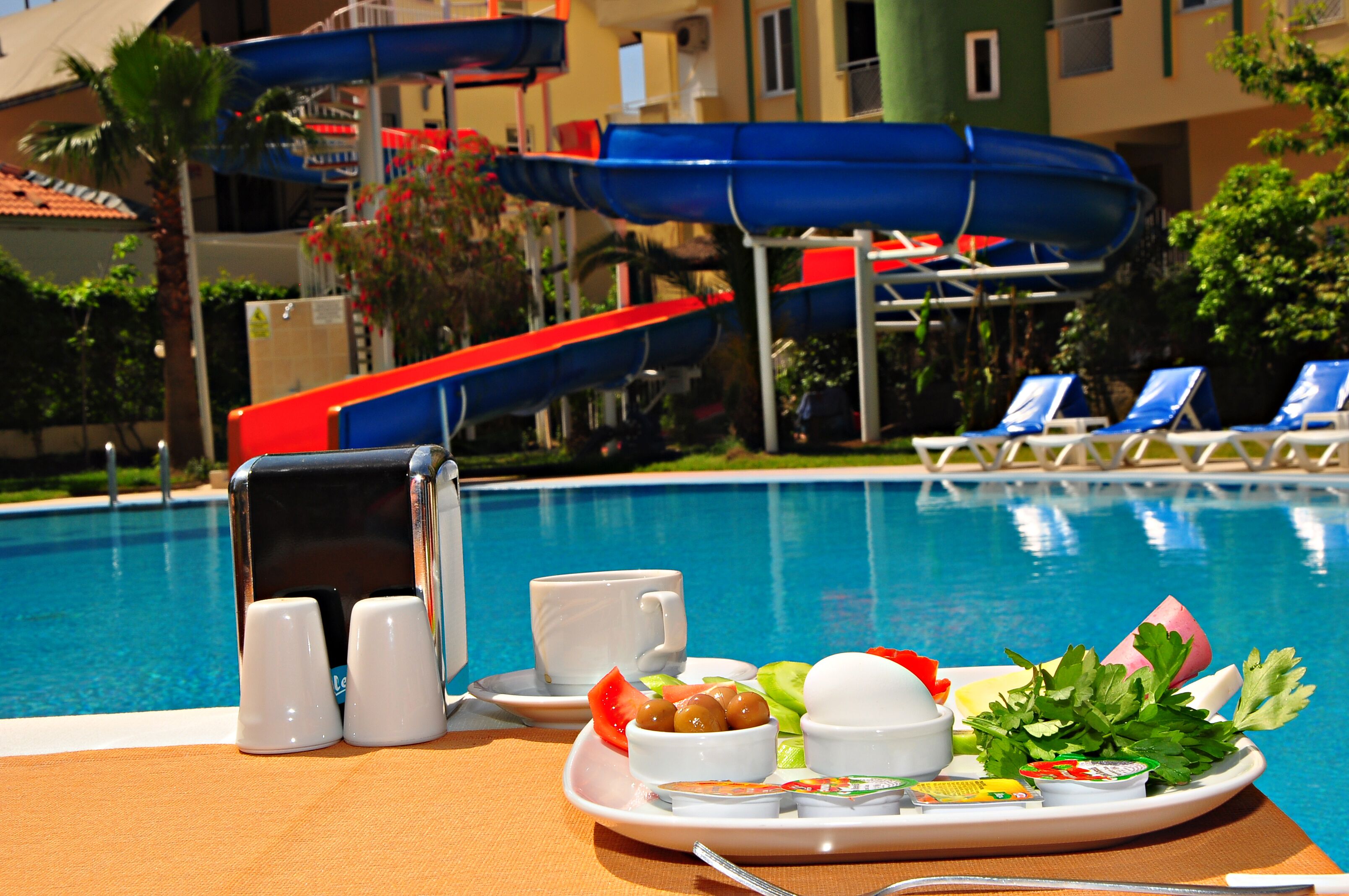 Ein Frühstücksbrettchen am Pool mit Getränken, Brot und Obst, im Hintergrund eine Wasserrutsche.