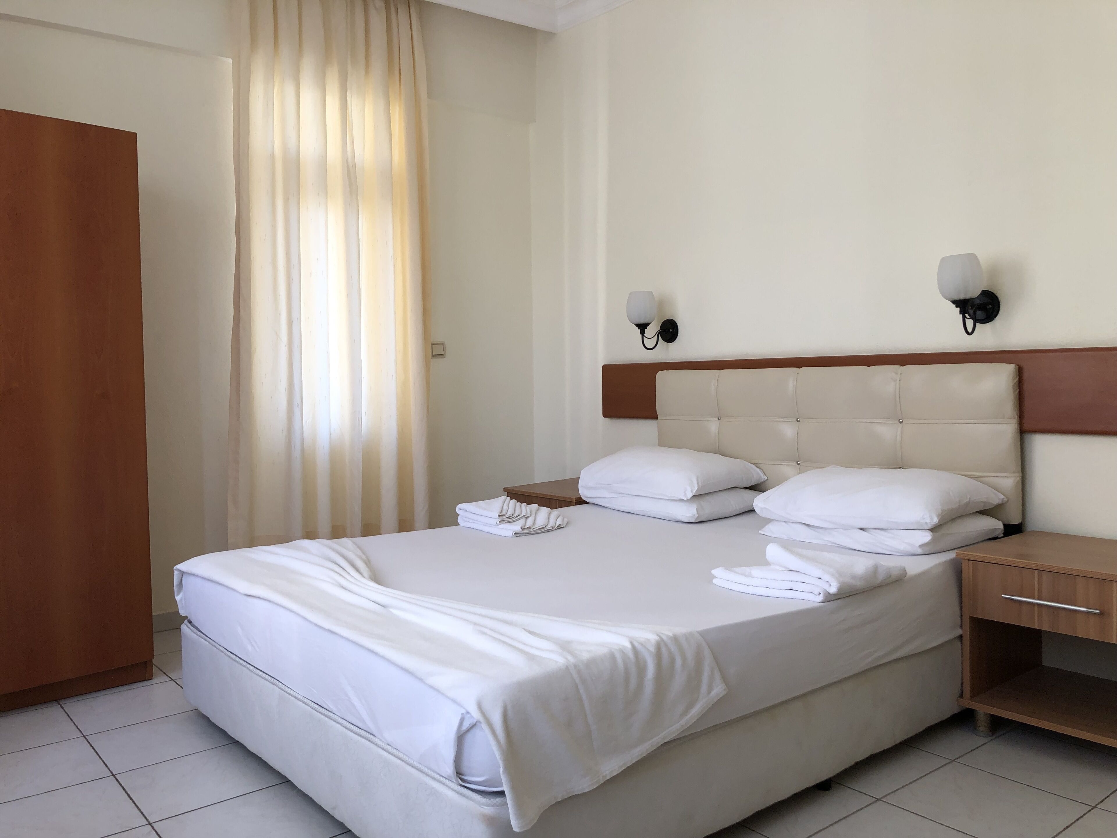 A hotel room with a bed, two bedside tables, a nightstand, a wooden wardrobe, and a window with curtains.