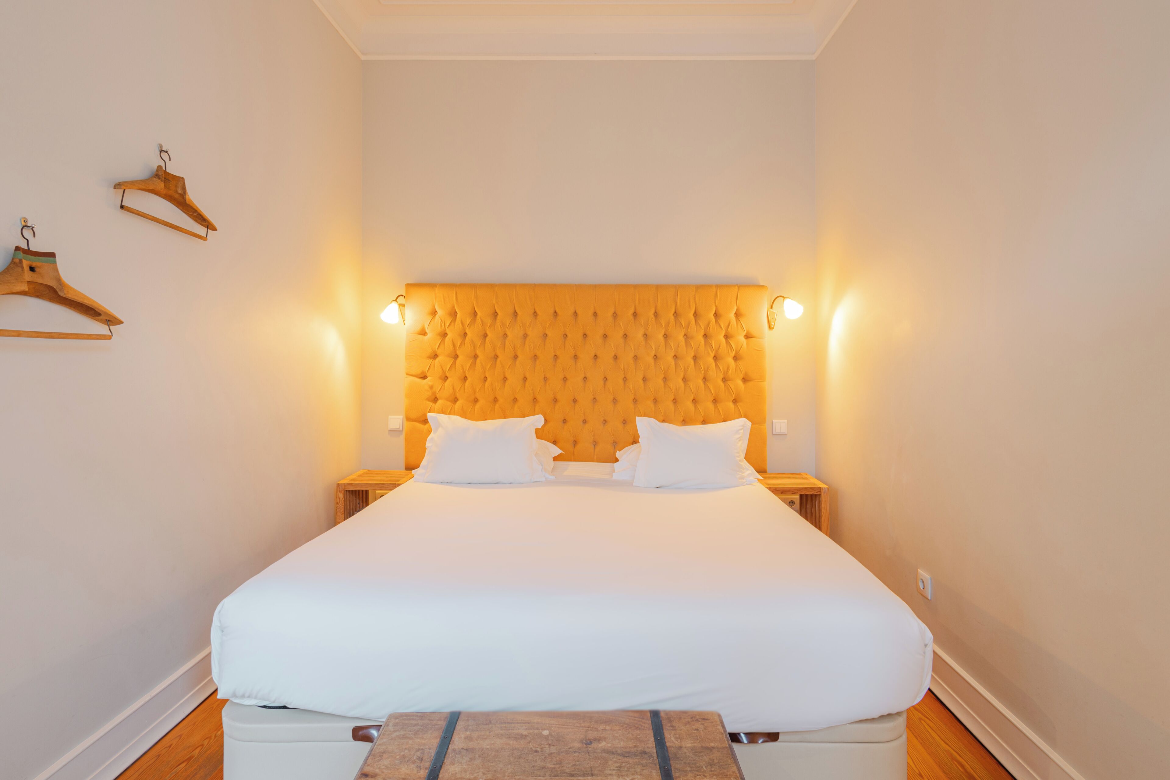 Una cama bien hecha con un cabecero capitonado, mesitas de noche y perchas de madera en la pared.