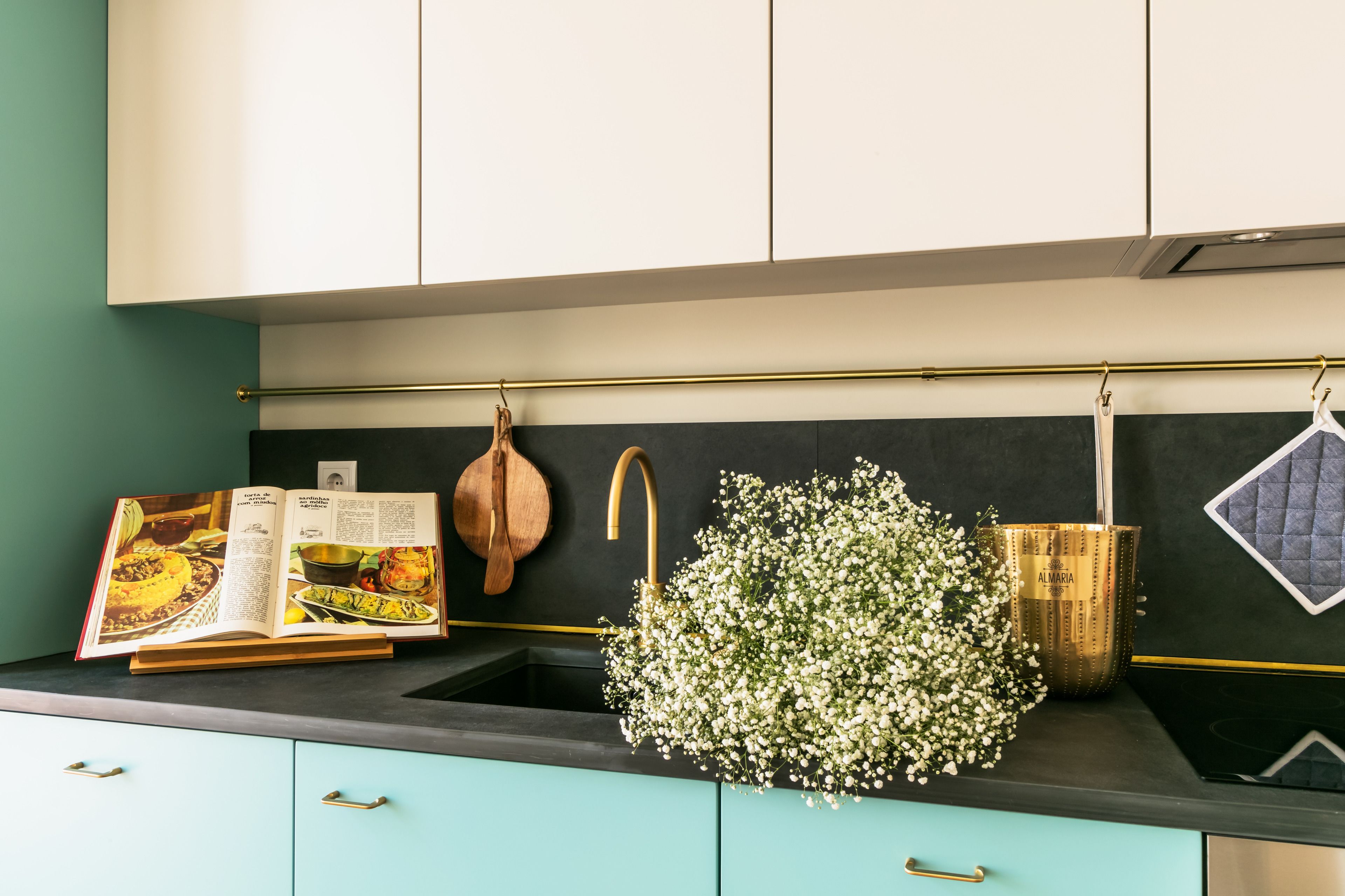 Una cocina con gabinetes color turquesa, salpicadero negro, un arreglo floral colgante, un libro de cocina y una olla de cobre.