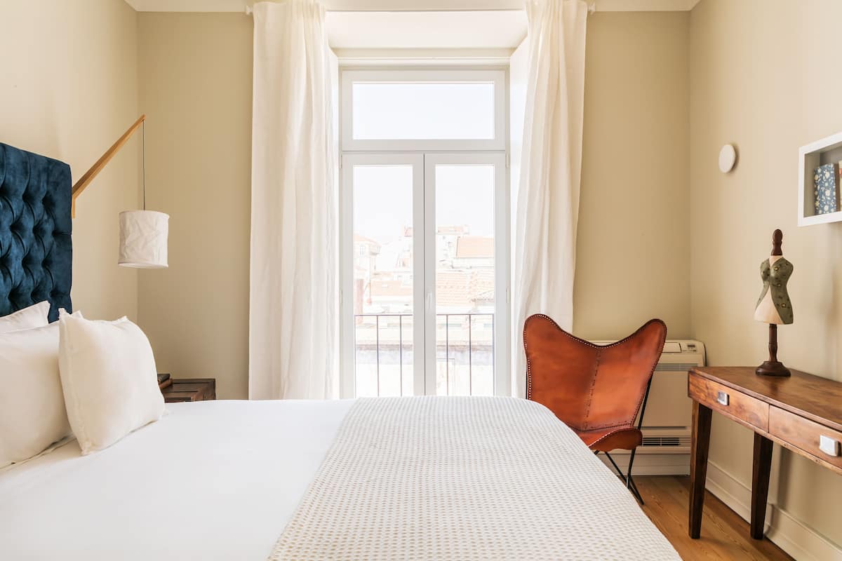 Una habitación de hotel con cama, una silla, un escritorio, una lámpara y una ventana con cortinas.