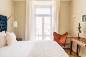 Quarto de hotel com cama, cadeira, escrivaninha, luminária e janela com cortinas.