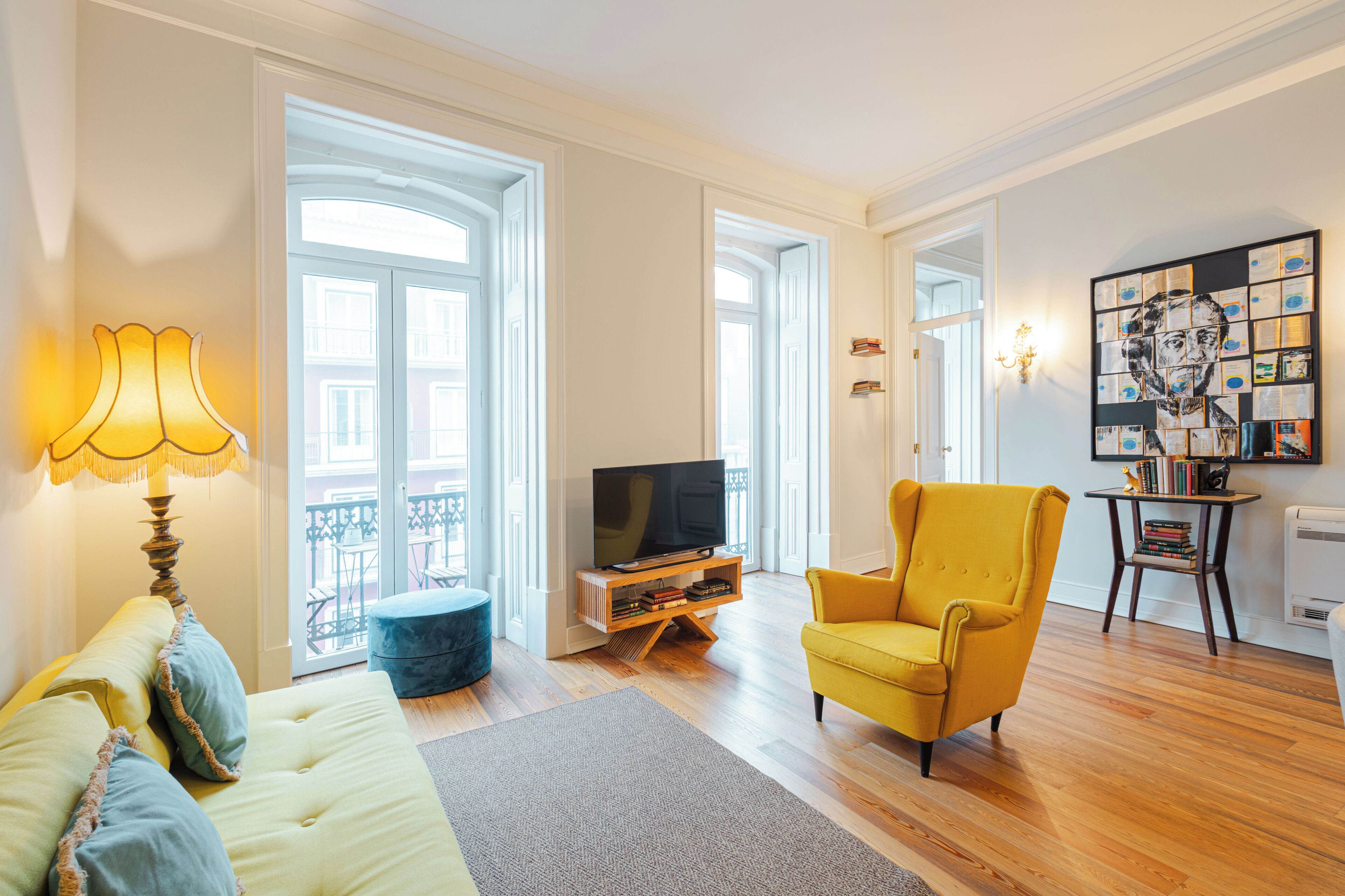 Una sala de estar con un sofá, un sillón amarillo, un televisor sobre un mueble de madera y una pared con una obra de arte enmarcada.