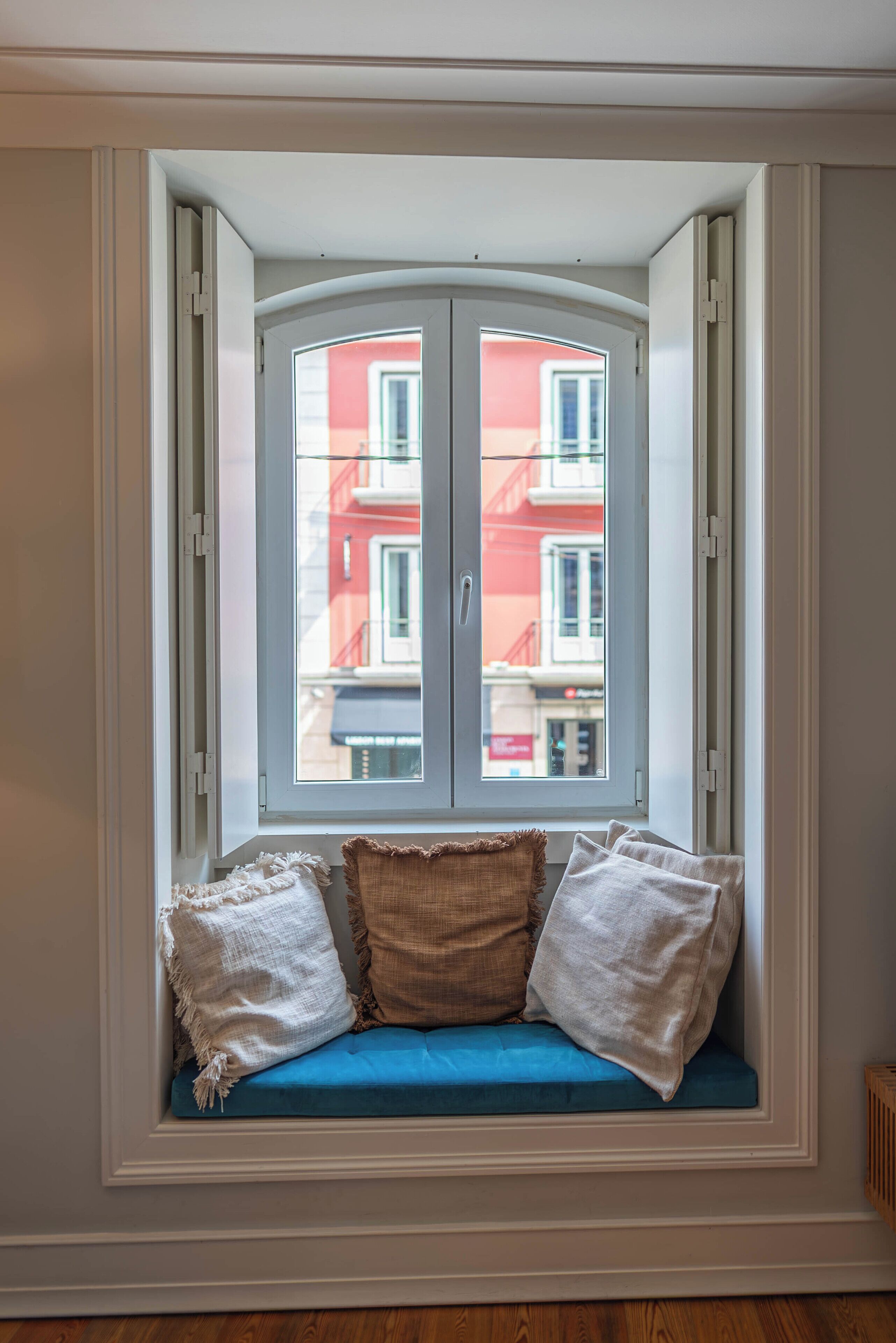 A bay window with a view of buildings, a blue cushioned seat, and two pillows.