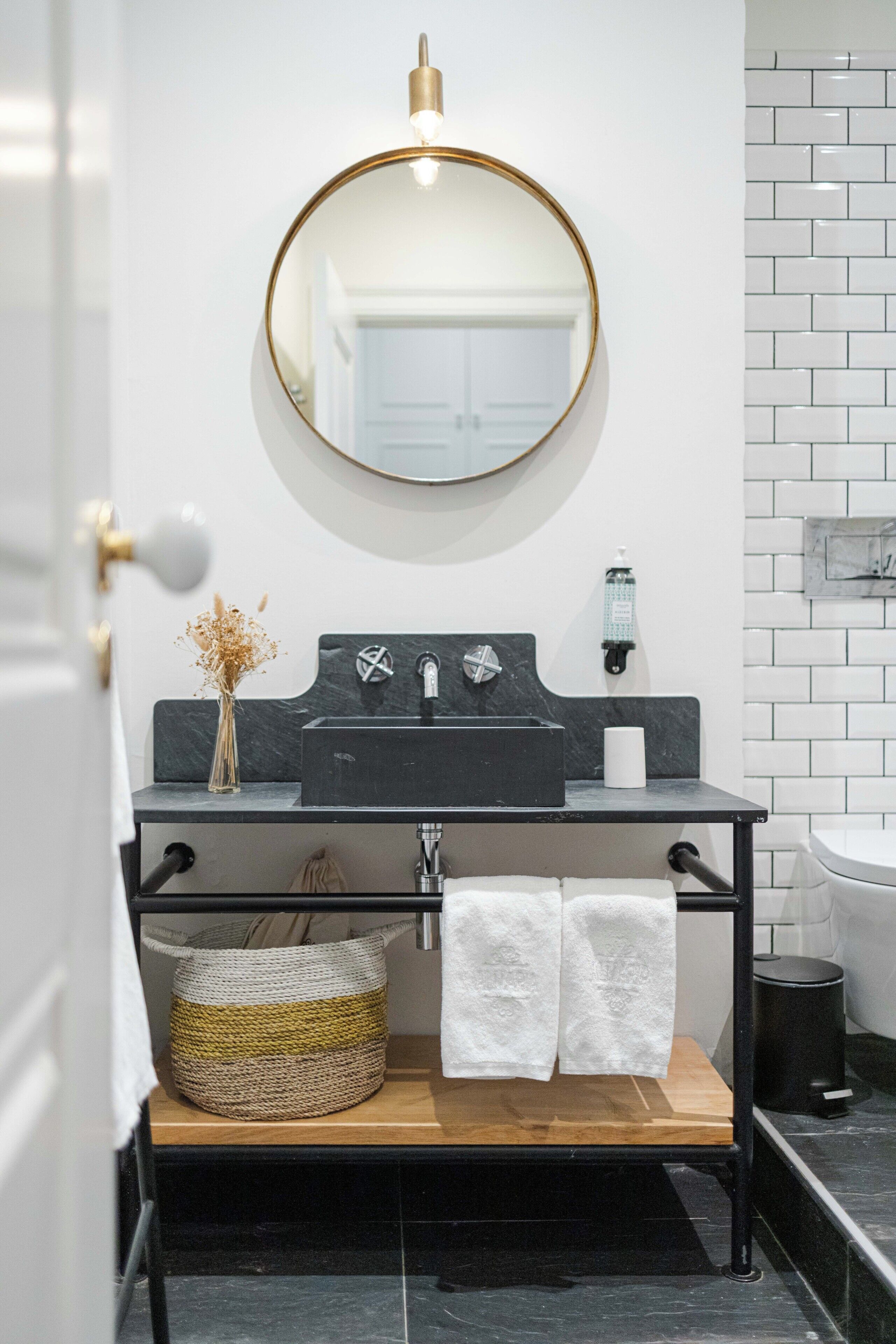A bathroom with a round mirror, black sink, white tiles, a wooden shelf, and a black trash bin.