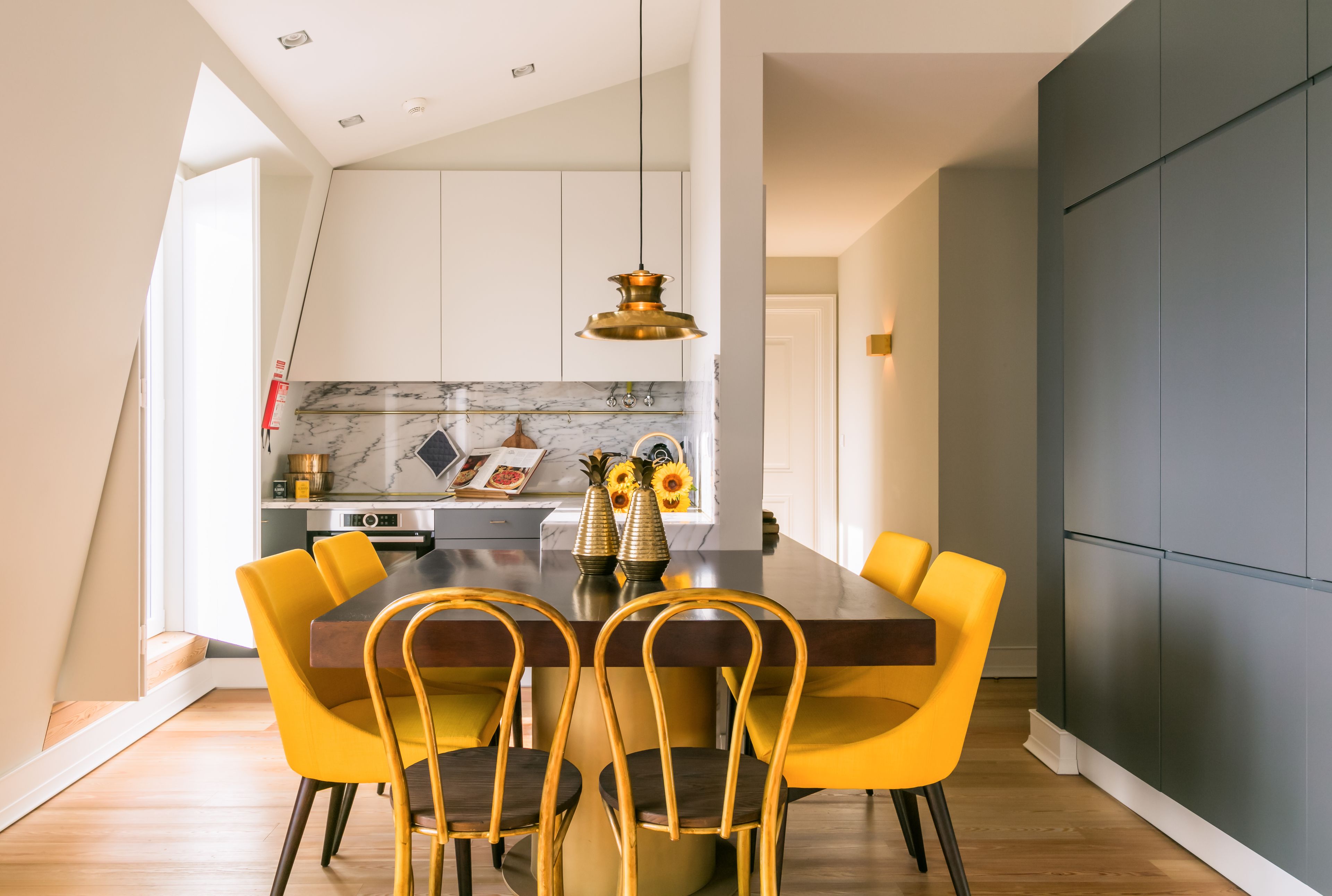 Uma área de refeições com uma mesa de madeira e cadeiras amarelas, uma cozinha ao fundo e uma luminária pendente moderna.