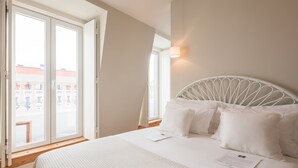 Un dormitorio con cama, ropa de cama blanca, ventana con vista a edificios y cabecera blanca.