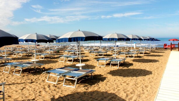 A beach with rows of sun loungers and umbrellas, a wooden walkway, and the ocean in the background. - Hotel Ariston (Grosseto)