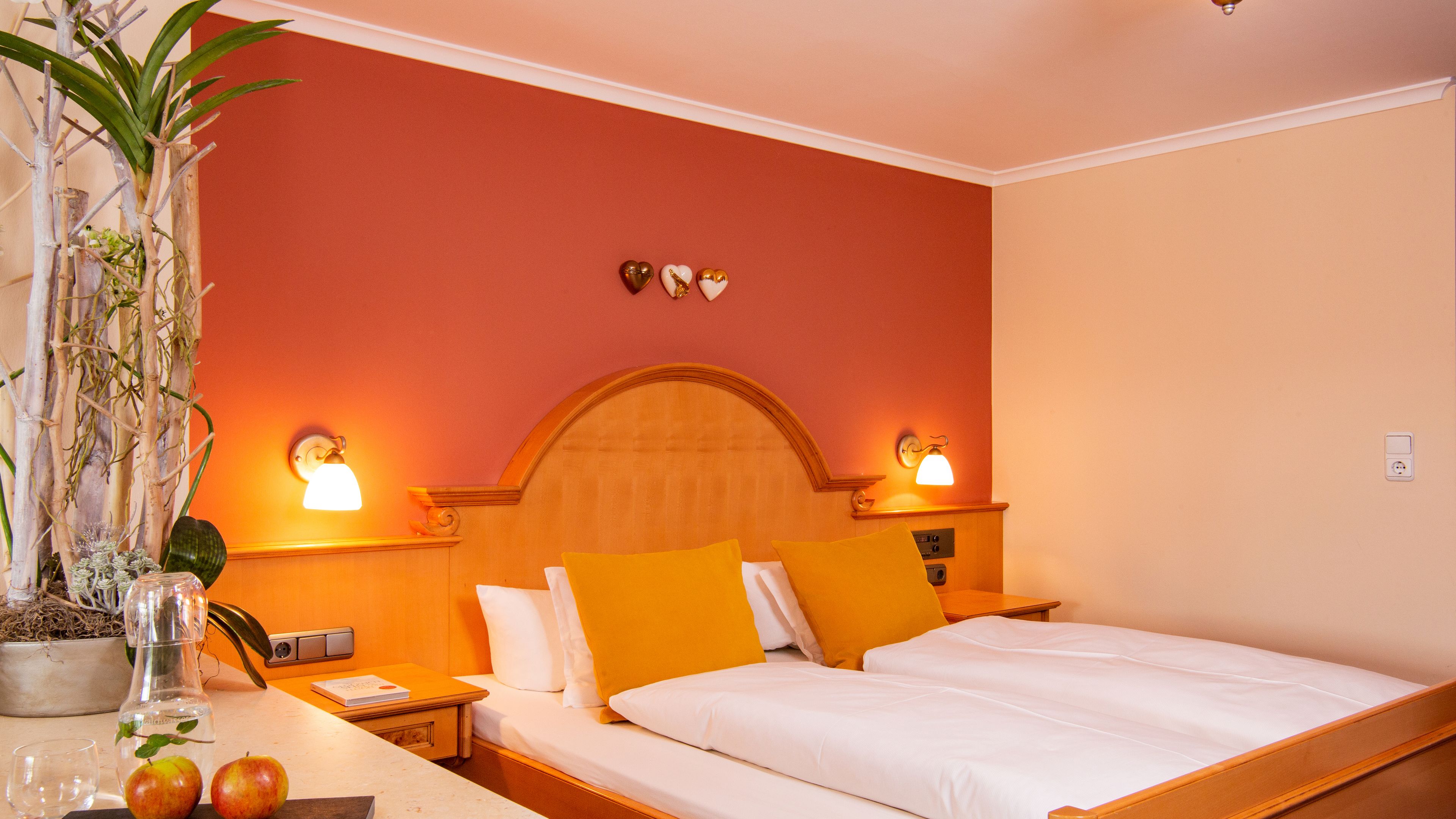 A hotel room with a bed, bedside tables, a nightstand, a potted plant, and wall-mounted lamps.