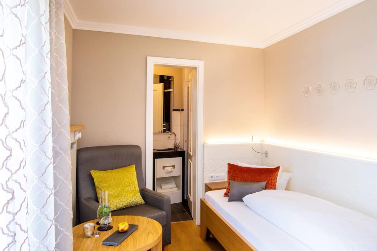 Una habitación de hotel con una cama, una silla con un cojín amarillo, una mesita con un vaso y una mesita de noche con una lámpara.
