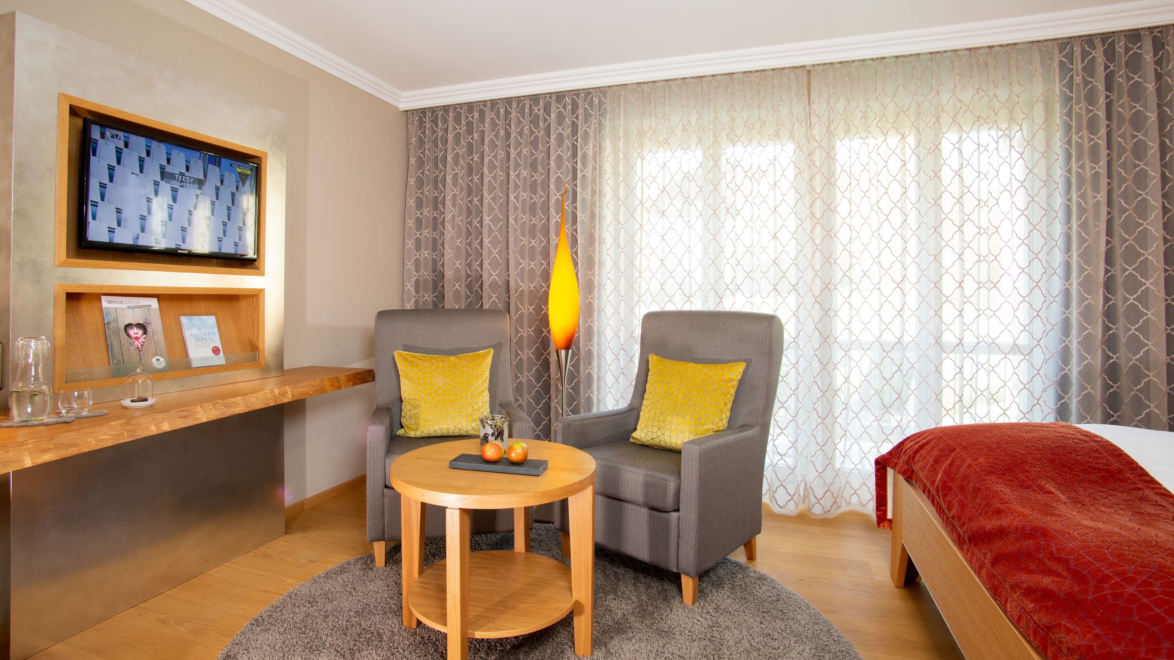 A hotel room with two grey armchairs, a small wooden table, a TV mounted on the wall, and a bed with a red blanket.