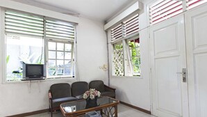 Una habitación con una televisión sobre un pedestal, dos sillas, una mesa de centro de vidrio con revistas y una puerta con marco blanco.