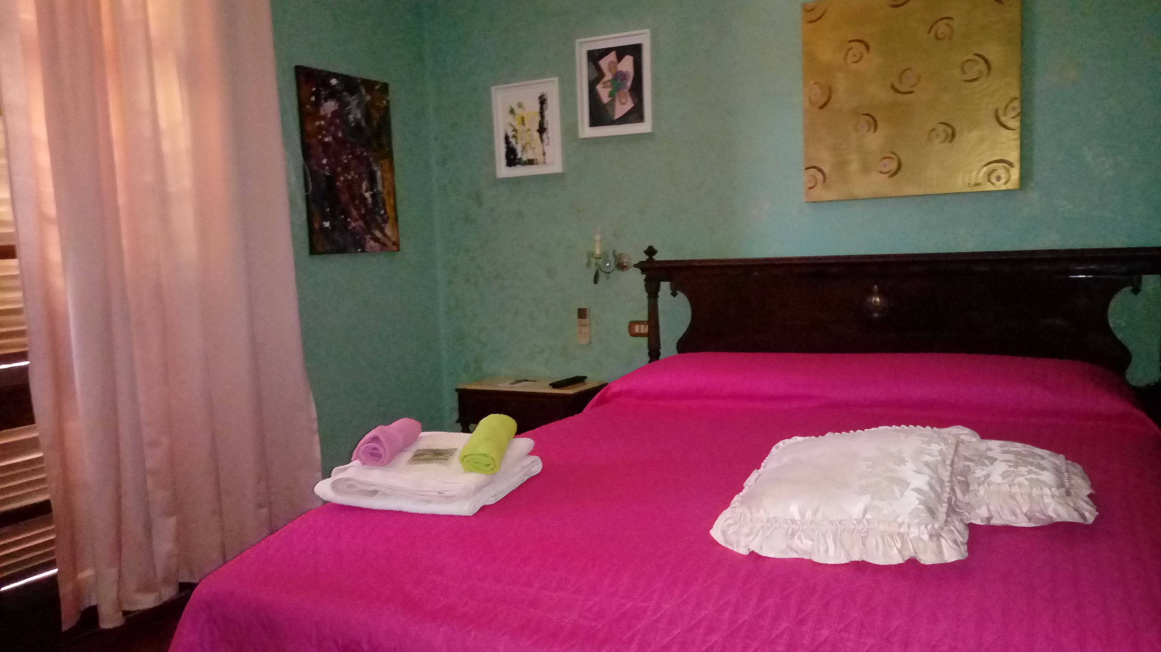 A room with a bed covered in a bright pink blanket, a white pillow, and a rolled towel on a tray. There are two framed pictures on the wall and a patterned curtain.