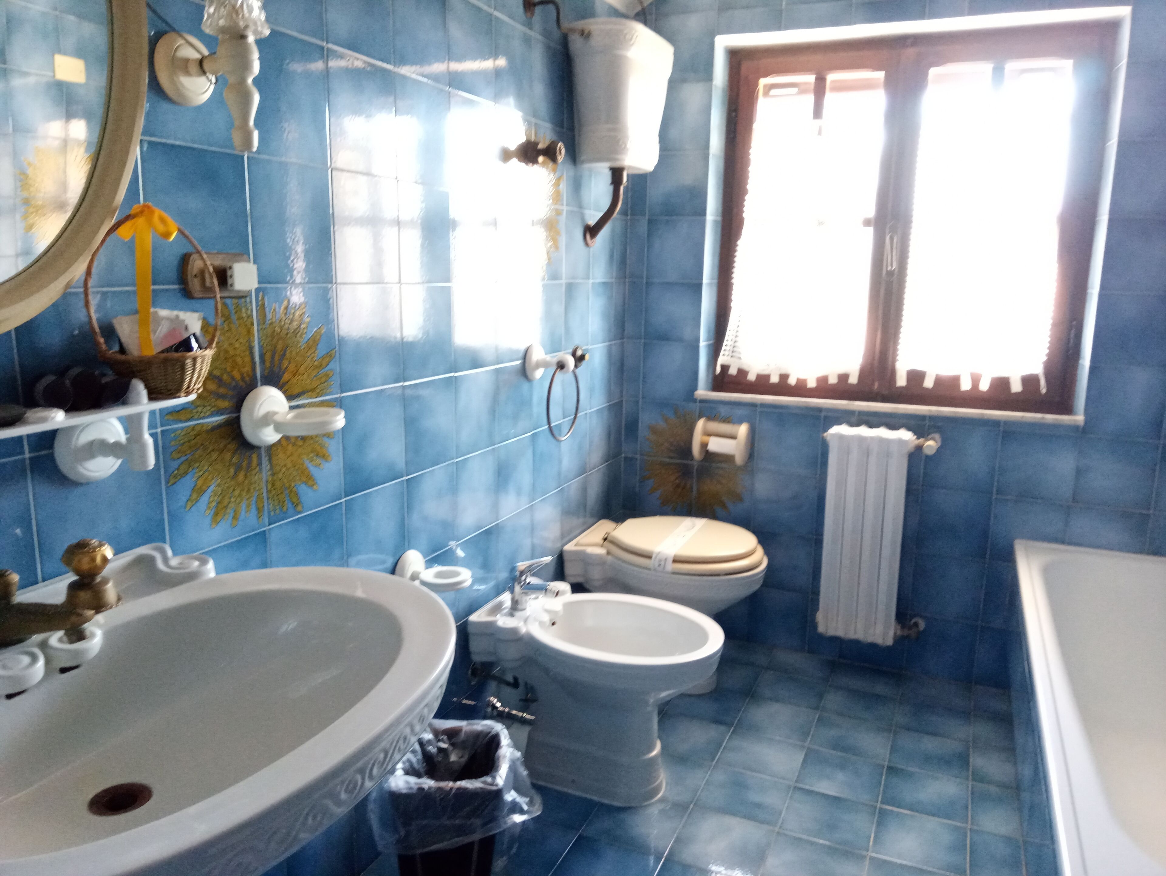 A bathroom with a large sink, toilet, and bidet. There is a window with curtains and a towel hanging on the wall.