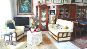 Uma sala de estar com sofá, poltronas, televisão, aparador e um quadro na parede.
