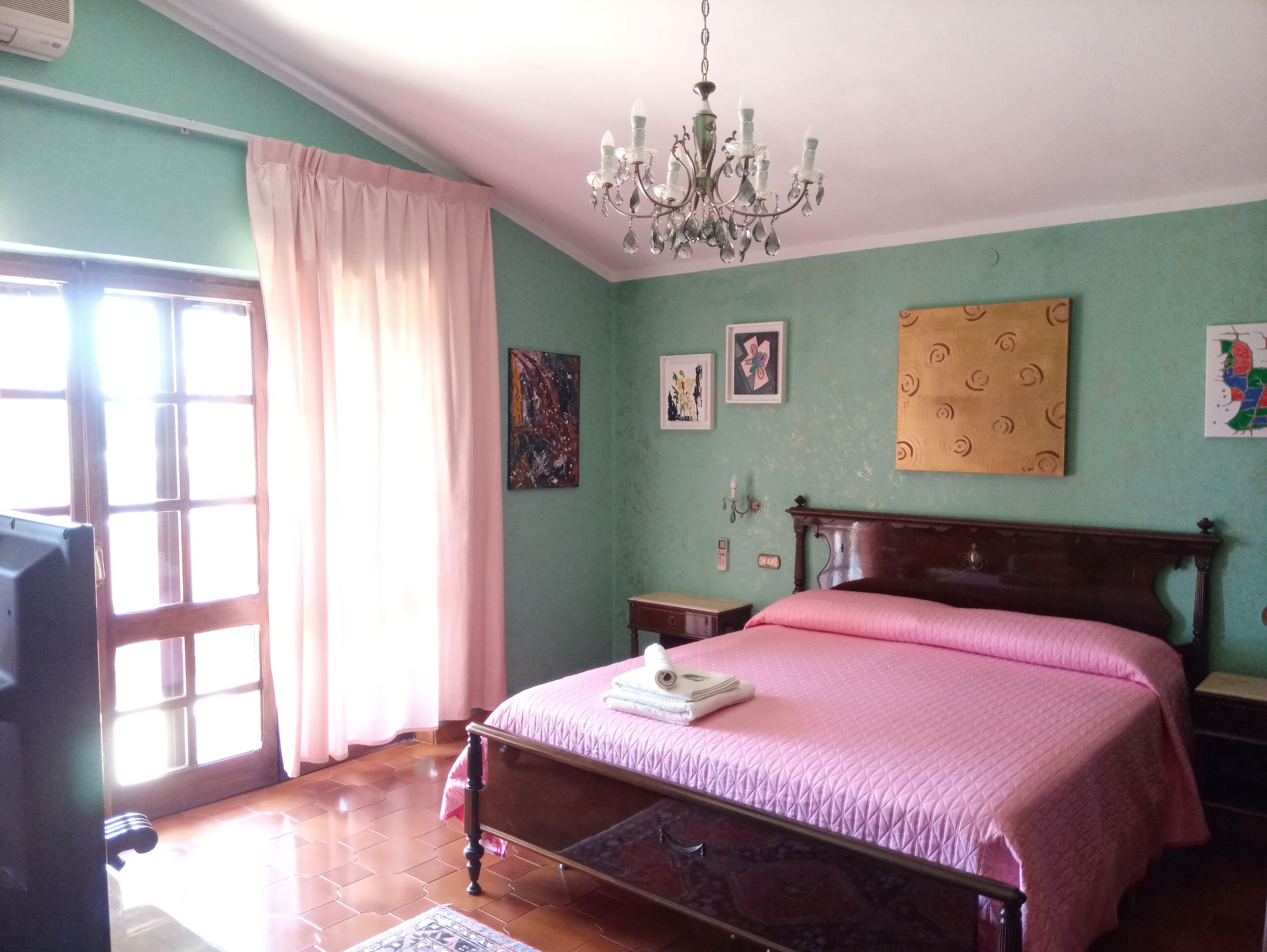 A bedroom with a bed, a chandelier, a window with curtains, and wall art.