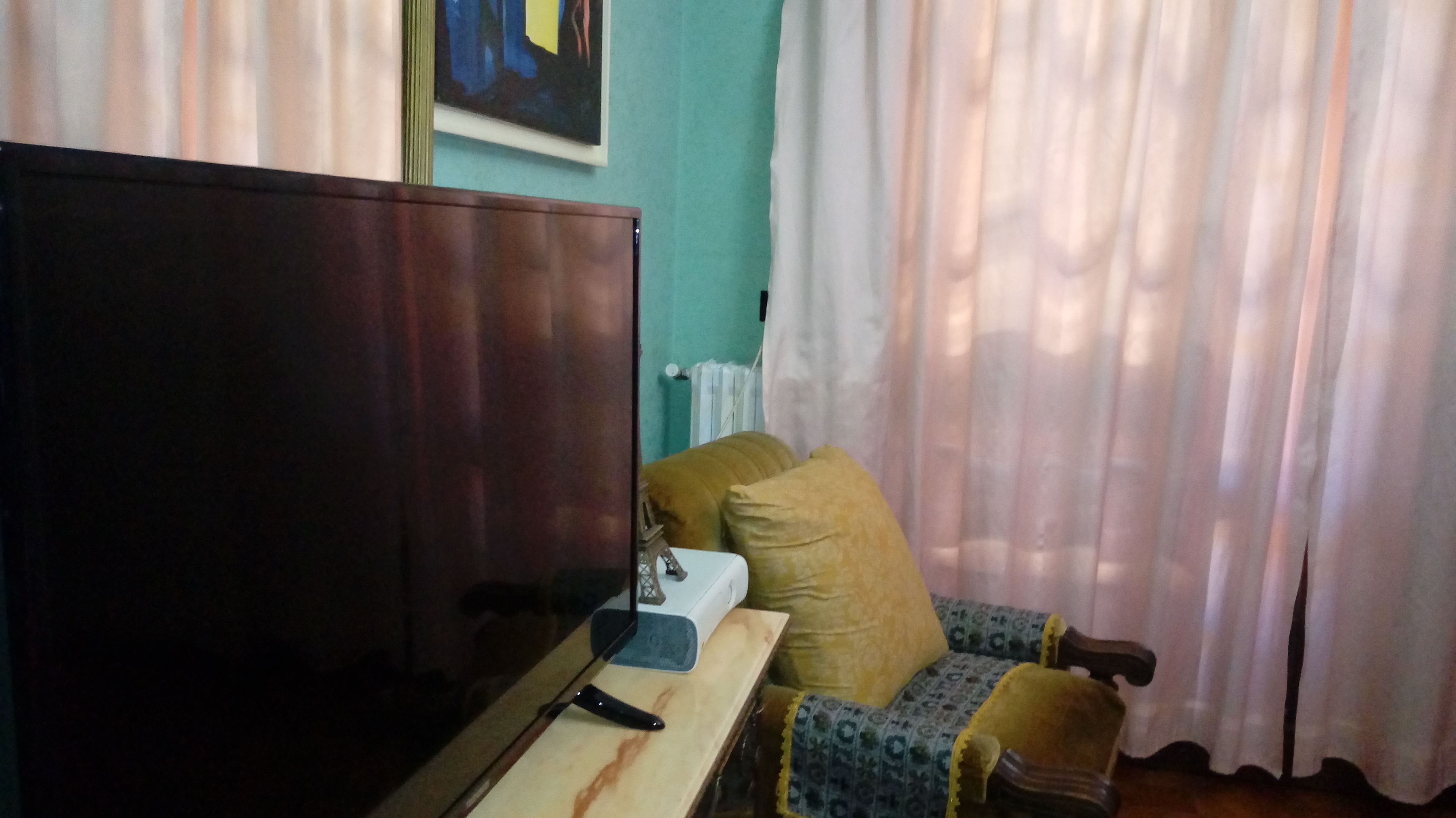 A room with a dark wooden wardrobe, a small table with a remote control, a patterned armchair, and a radiator.