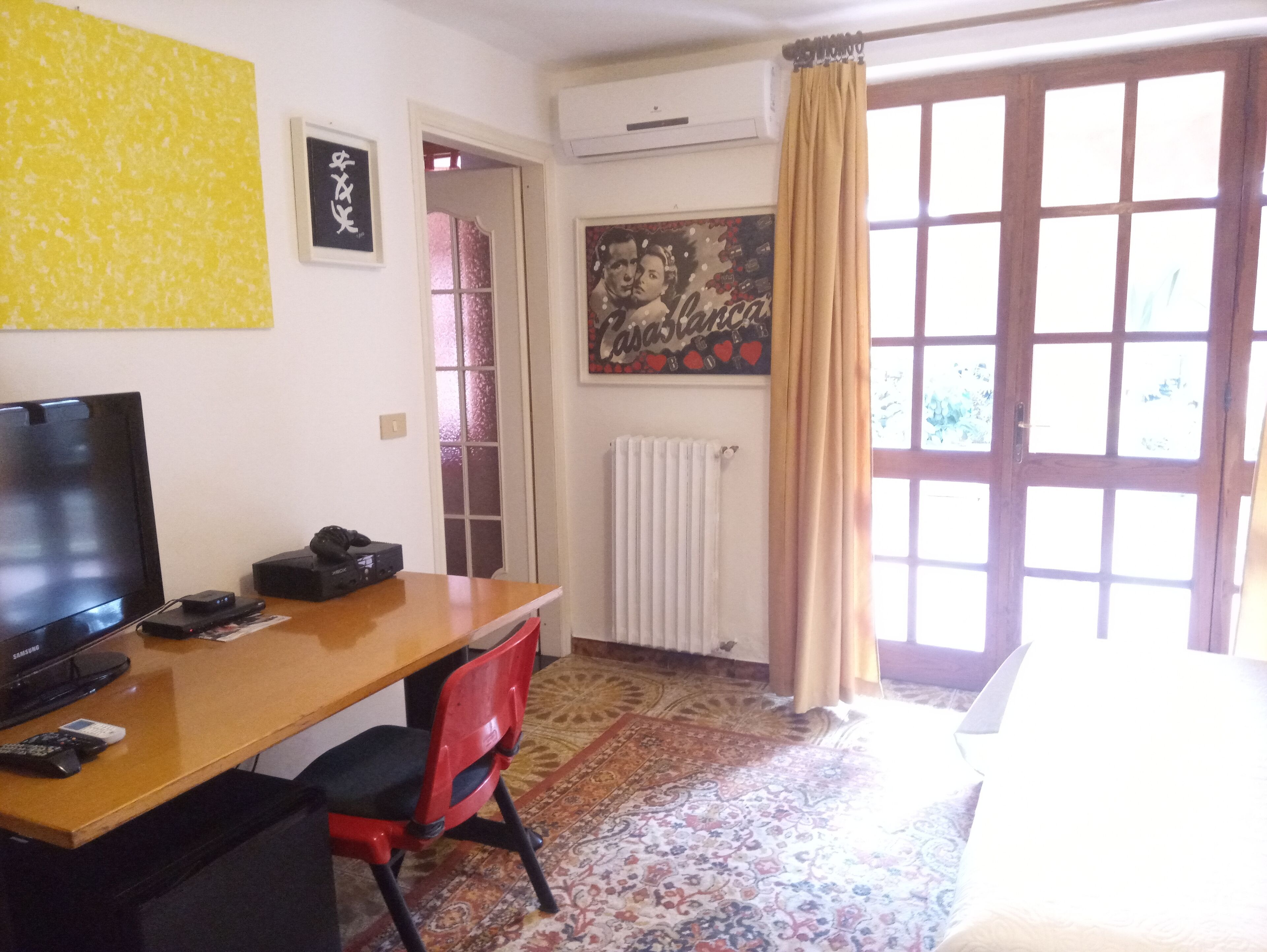Ein Zimmer mit Schreibtisch aus Holz, einem Fernseher, einem roten Sessel, einer Heizung, einem gemusterten Teppich und einem großen Fenster mit Vorhängen.