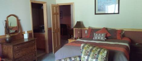 Un dormitorio con cama, una silla, cómoda y espejo. Hay una puerta que da a otra habitación.