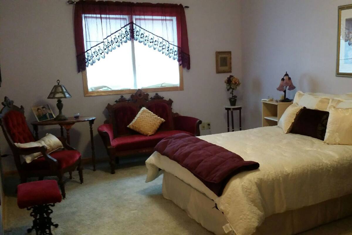 Een slaapkamer met een groot bed, een rode fauteuil, een houten stoel, een kleine tafel met een lamp en een raam met kantgordijnen.