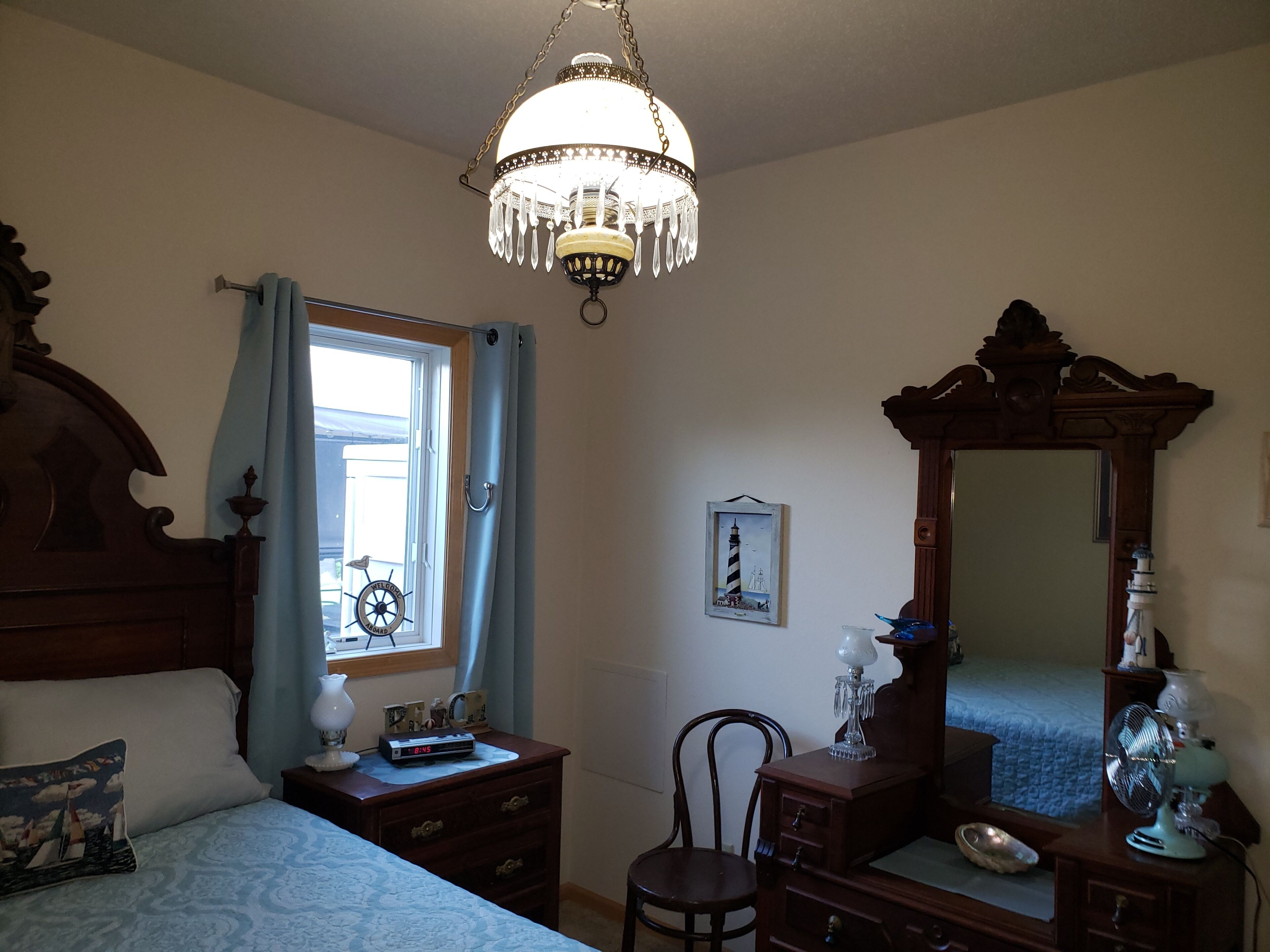 Un dormitorio con cama de madera, mesita de noche, cómoda, silla, espejo grande, candelabro y ventana con cortinas.