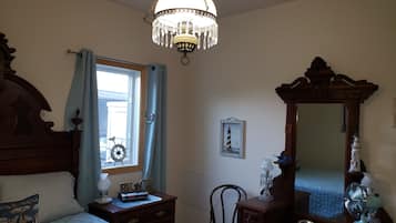 Un dormitorio con cama de madera, mesita de noche, cómoda, silla, espejo grande, candelabro y ventana con cortinas.