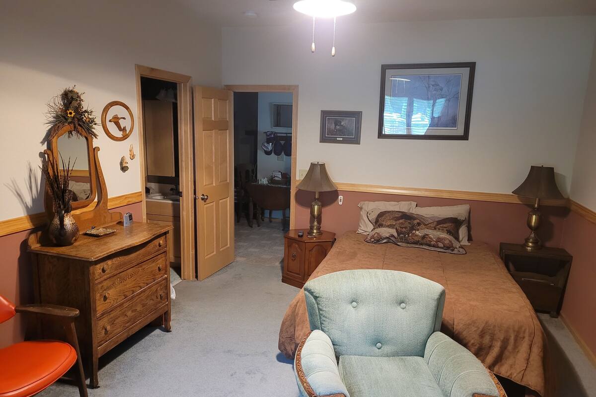 Een slaapkamer met een bed, een stoel, een dressoir, een spiegel en een deur naar een badkamer.