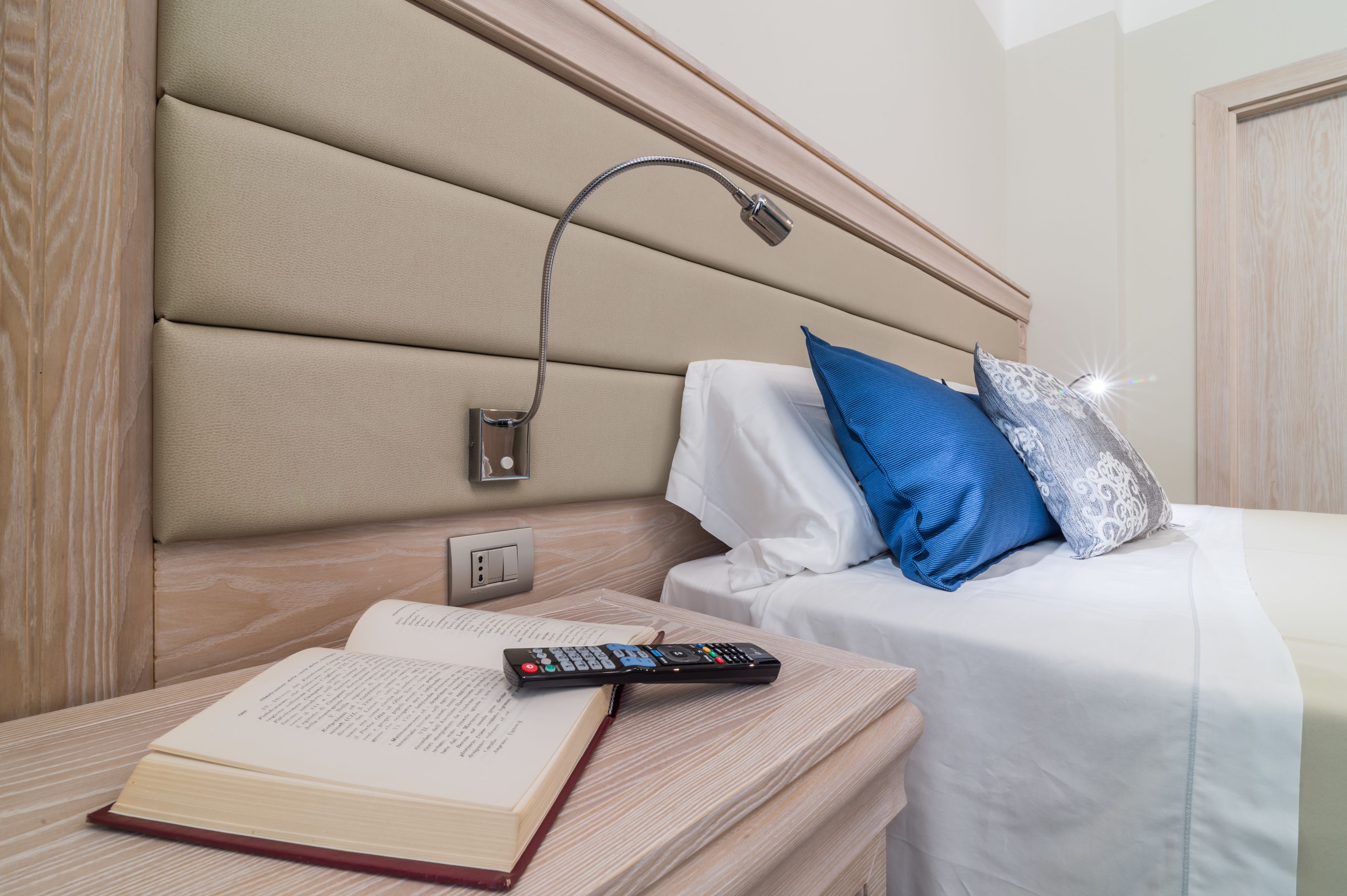 A neatly made bed with a headboard, a bedside table with a book and remote control, and a wall-mounted night light.