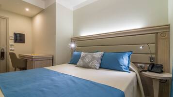 A hotel room with a bed, a desk, a chair, a bedside table with a phone, and a wall-mounted TV.