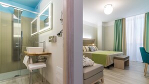 Una habitación de hotel moderna con cabina de ducha de vidrio, una mesita de noche con lavabo y una cama con colcha verde, además de una silla.