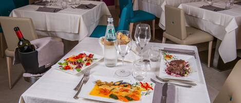 Une scène de restaurant avec des tables dressées pour le dîner, où l’on voit des assiettes remplies de nourriture, des verres à vin et des couverts.