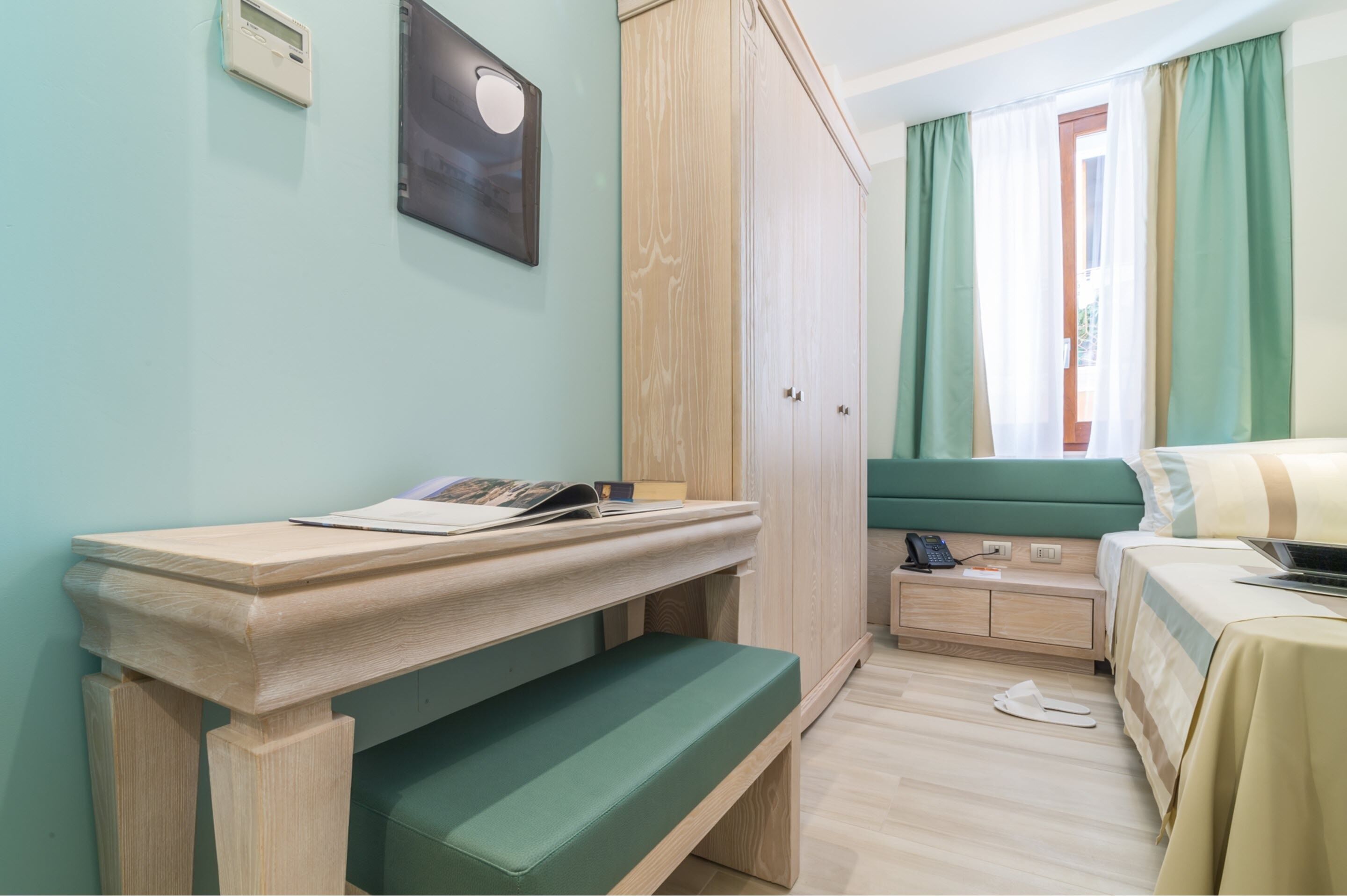 A room with a wooden desk, a green bench, a bed with a green headboard, a wardrobe, and a window with curtains.