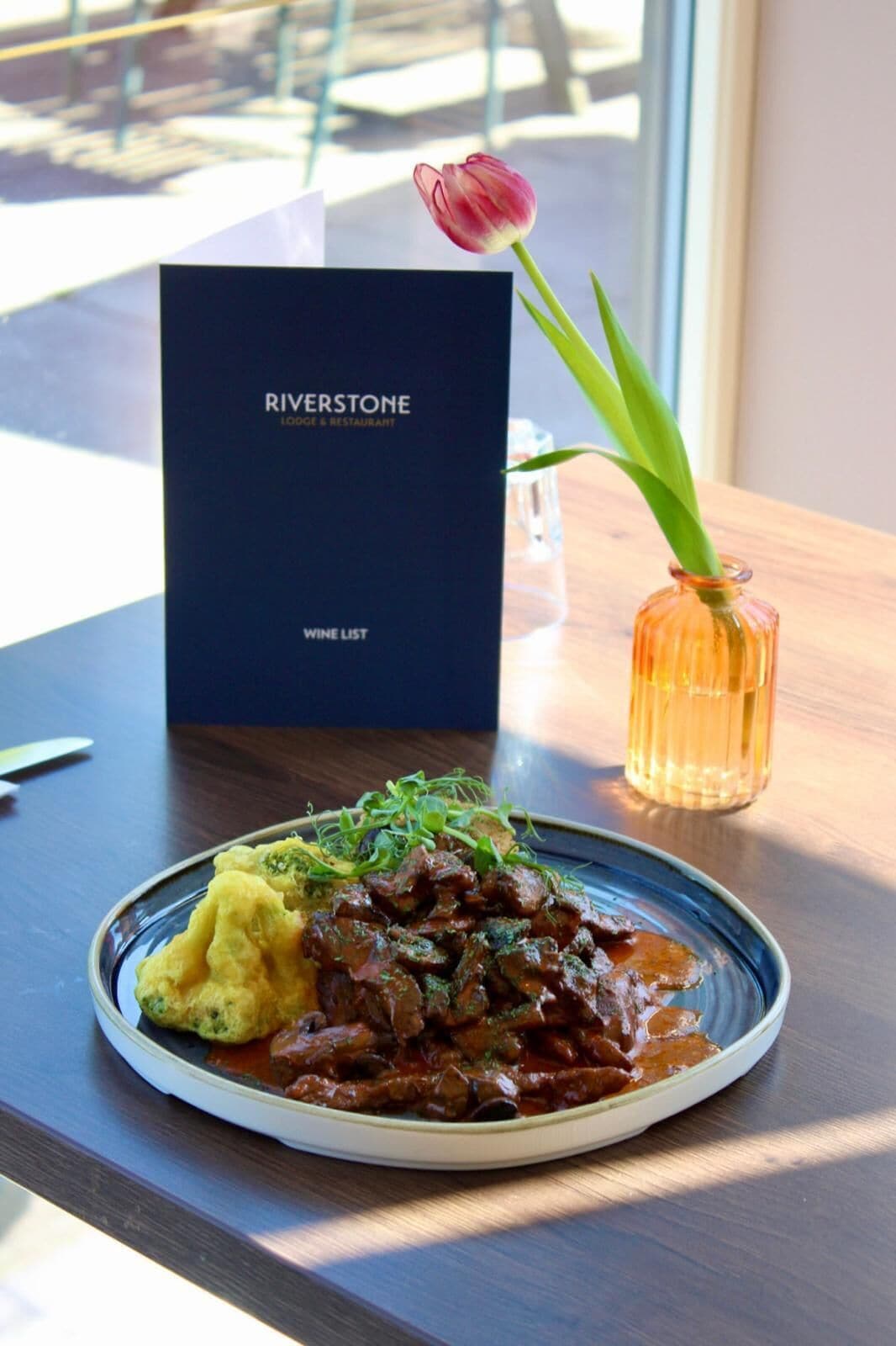 En tallrik med mat, en meny med titeln ”RIVERSSTONE LODGE & RESTAURANT” och en vas med en blomma.