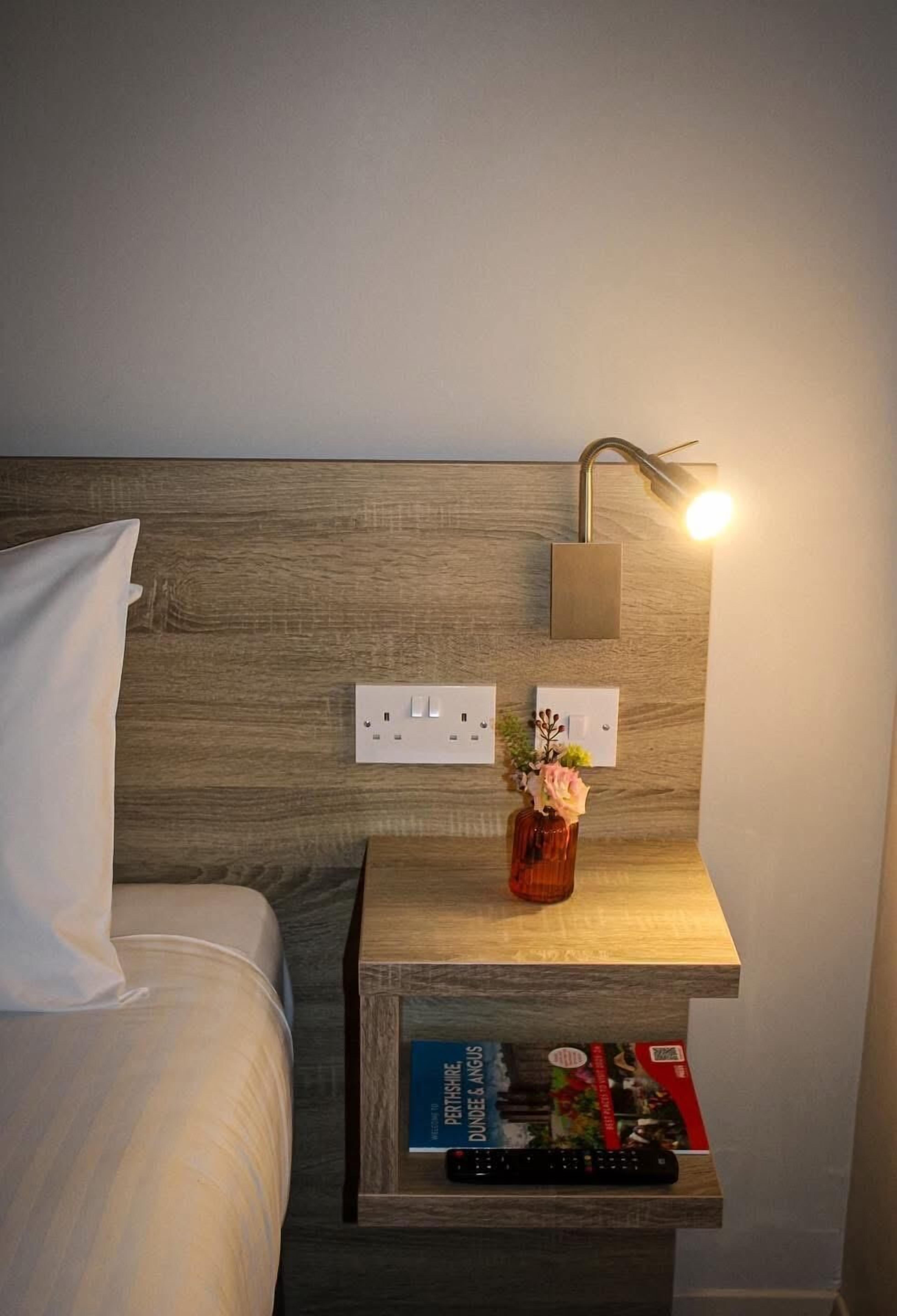 A hotel room with a wooden headboard, a bedside table with a remote control, a vase with flowers, and a book.