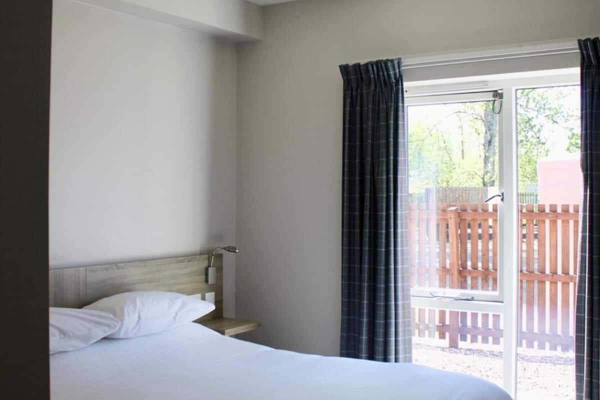 A bedroom with a bed, a window with curtains, a ceiling light, and a view of a wooden deck outside.