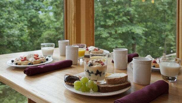 Mesa de café da manhã com diversos alimentos e bebidas, com vista para árvores através de uma janela.