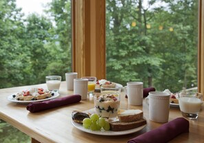 A breakfast table with various food items and drinks, set against a backdrop of trees through a window. - Fenton Inn (Roseland)