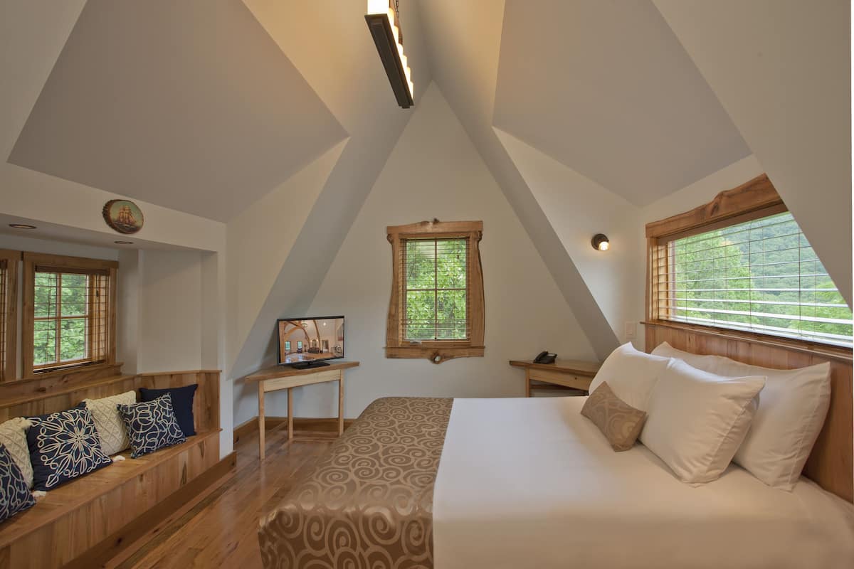 A bedroom with a sloped ceiling, a bed with white bedding, a wooden bench with patterned cushions, a small table with a lamp, and a window with blinds.