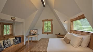 Une chambre avec un plafond en pente, un lit avec une literie blanche, un banc en bois avec des coussins à motifs, une petite table avec une lampe et une fenêtre avec des stores.