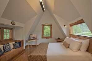 A bedroom with a sloped ceiling, a bed with white bedding, a wooden bench with patterned cushions, a small table with a lamp, and a window with blinds. - Fenton Inn (Roseland)