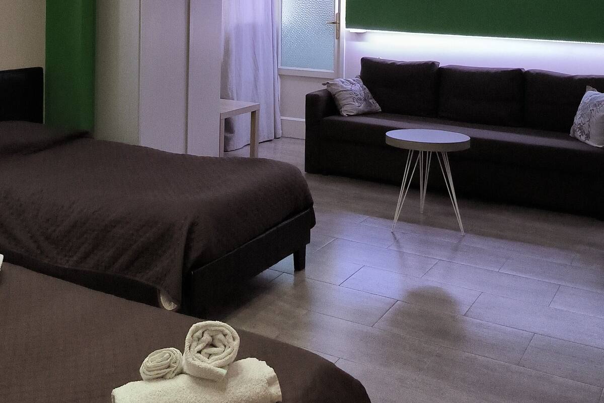 Une chambre d’hôtel avec deux lits, un canapé, une petite table, un téléviseur fixé au mur et une fenêtre avec des rideaux.