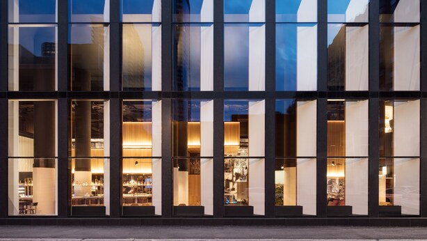 Un edificio moderno con amplios ventanales que reflejan el cielo y edificios cercanos.