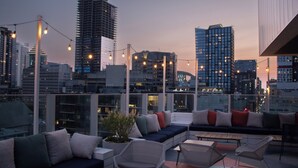 Terraza en la azotea con un área de estar moderna, que incluye sofás y sillones, con vistas a un paisaje urbano al atardecer.