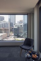 Una habitación de hotel moderna con un ventanal amplio que ofrece vistas a la ciudad, un sillón cómodo, una mesita con alimentos para el desayuno y una taza de café.